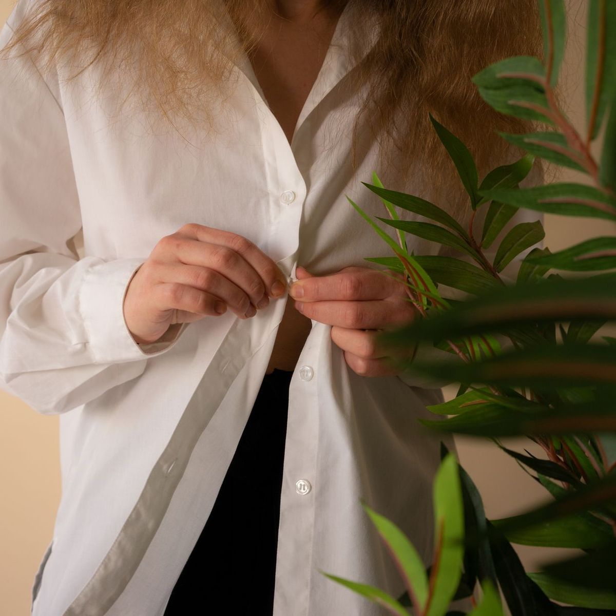 Person buttoning a loose white linen shirt beside a leafy plant